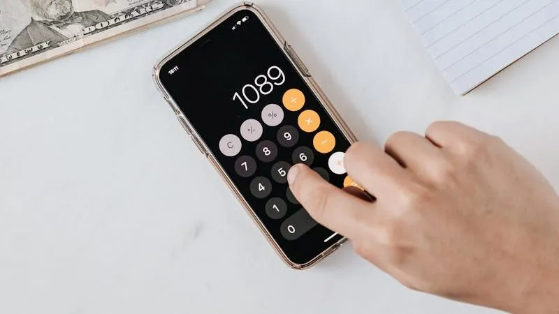A hand tapping a running total into a phone calculator app, beside banknotes and a lined notepad